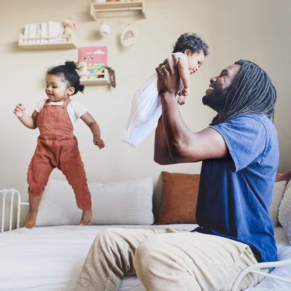 Father lifting smiling baby dressed in aden + anais swaddle in a bright nursery, toddler playing nearby.