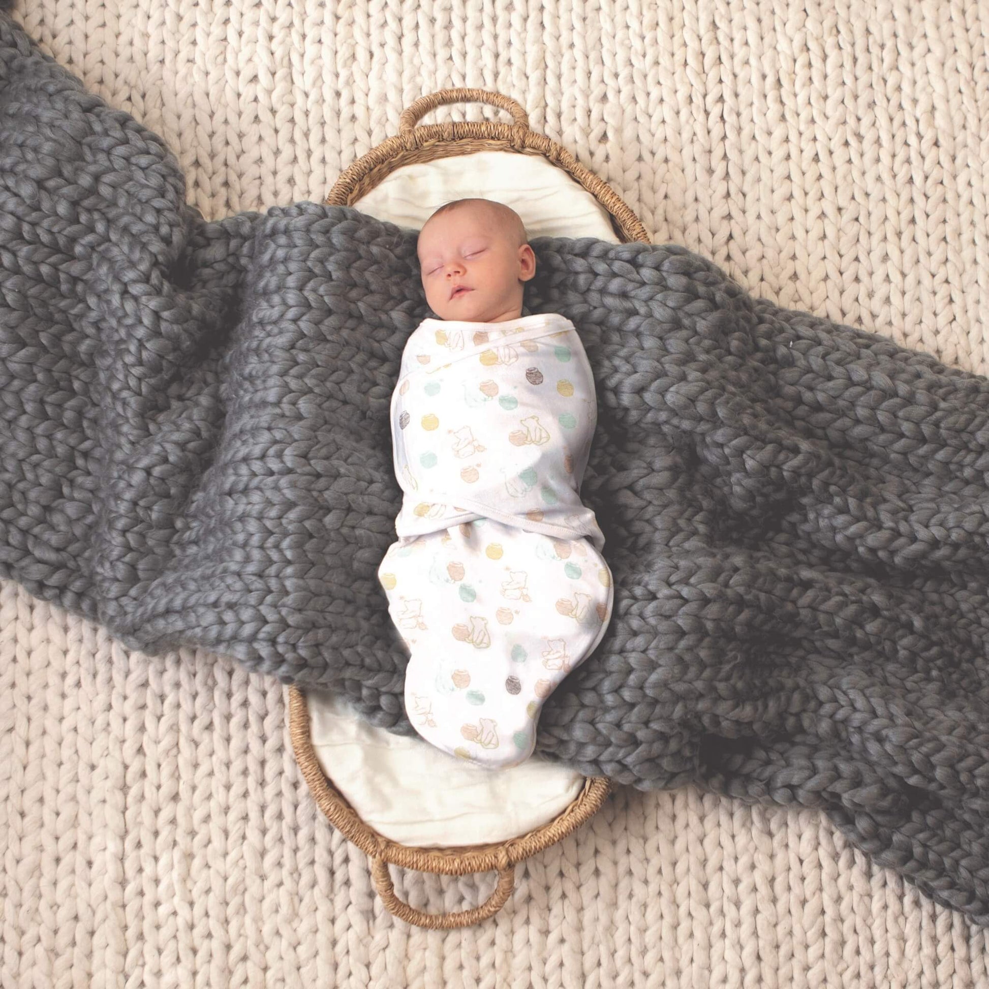 A baby is swaddled in a white wrap featuring pastel honey pots and outline drawings of Winnie the Pooh in various poses, lying in a basket with a thick grey knit blanket and cream textured rug underneath.