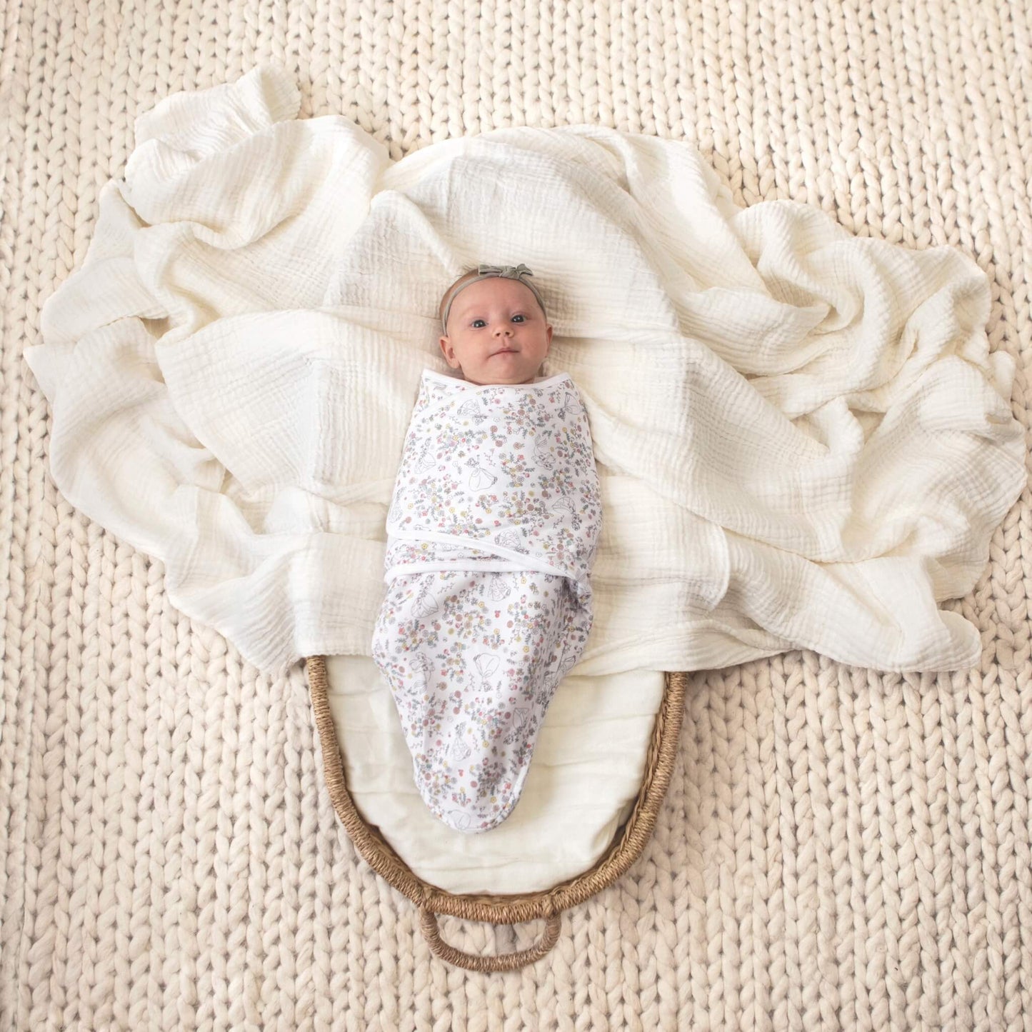 A baby lies swaddled in a white wrap with pastel floral and princess illustrations, placed in a woven basket lined with a soft white blanket on a textured cream rug.
