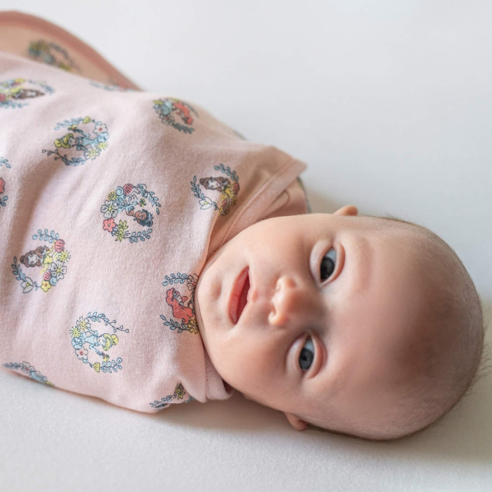 A baby lying on their side swaddled in a pink wrap printed with circular princess illustrations surrounded by small floral wreaths.