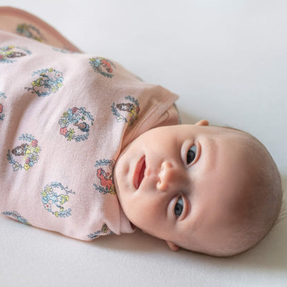 A baby lying on their side swaddled in a pink wrap printed with circular princess illustrations surrounded by small floral wreaths.