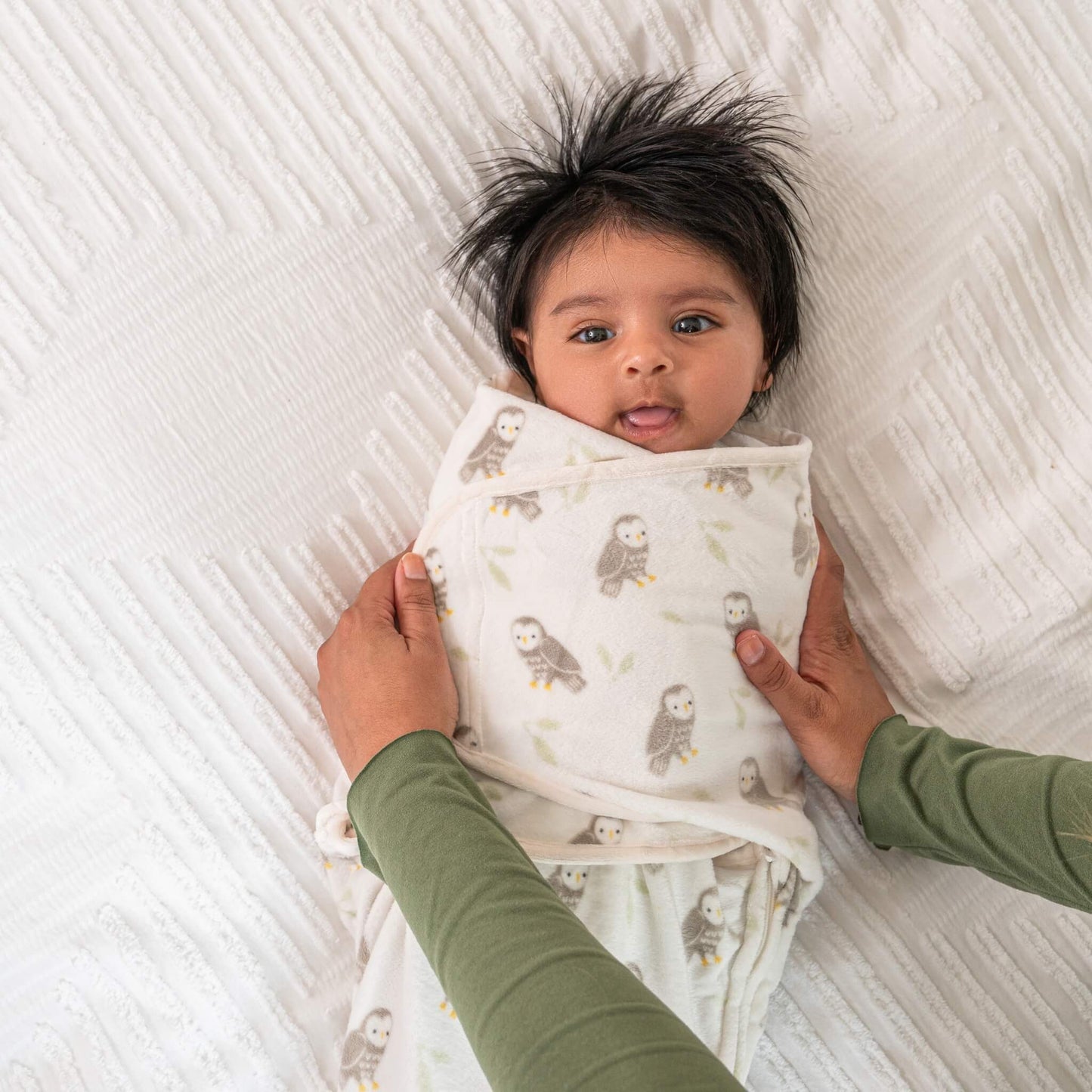 Mother holding baby swaddled in the owl print Rockland Marsh wrap, baby smiling and gazing up from a white textured surface.