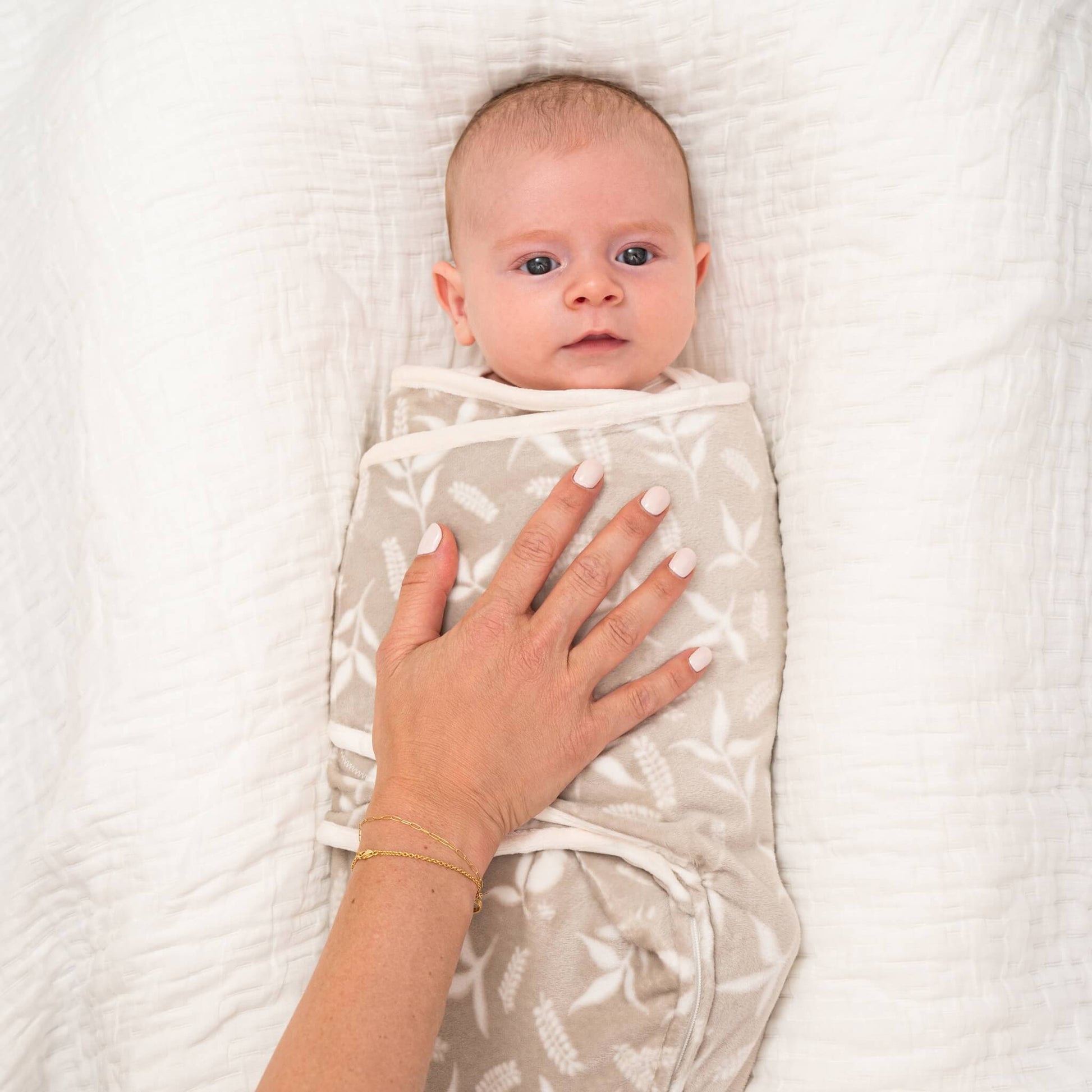 Close-up of mother’s hand gently resting on a baby swaddled in the beige Rockland Marsh velboa wrap, lying on a soft white blanket.