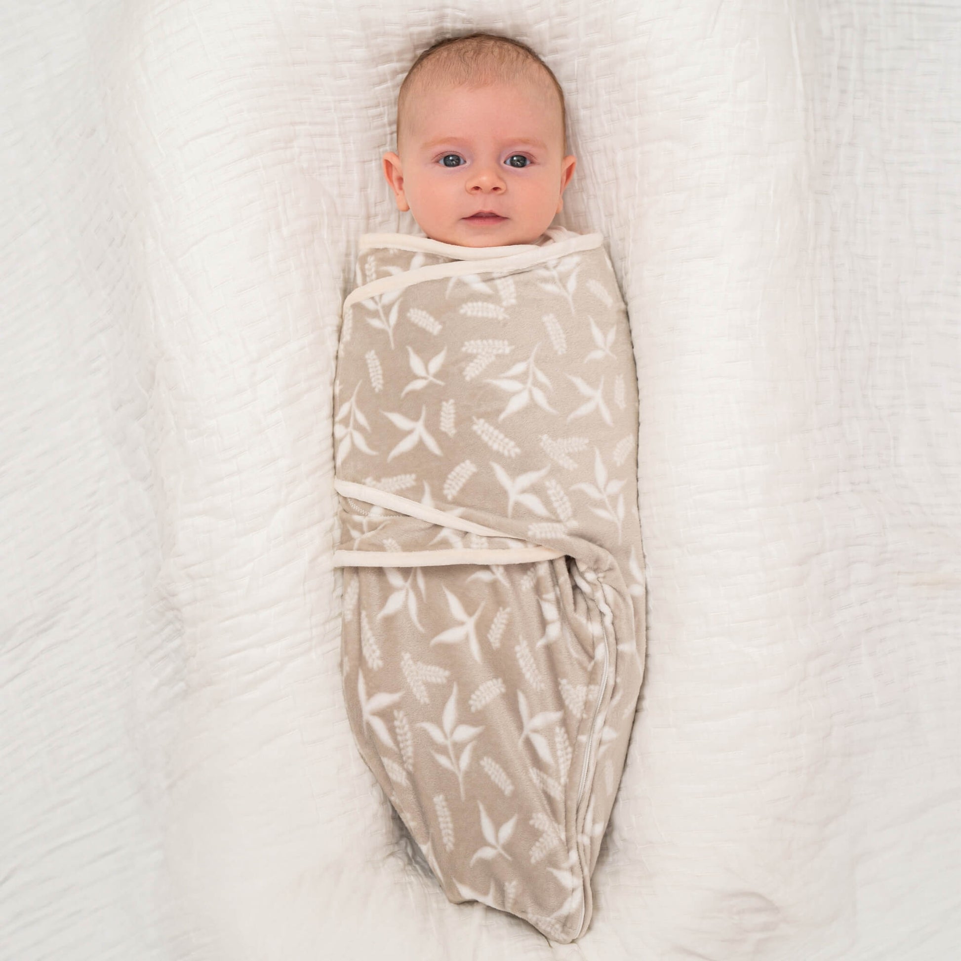 Baby wrapped in the Rockland Marsh beige swaddle, lying calmly on a plush white blanket with their arms tucked in.