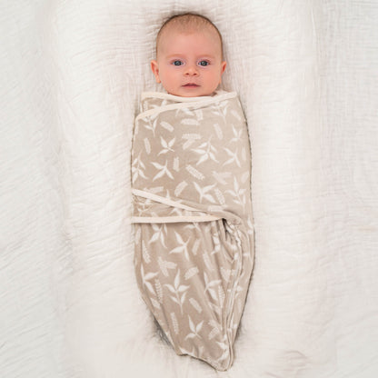 Baby wrapped in the Rockland Marsh beige swaddle, lying calmly on a plush white blanket with their arms tucked in.
