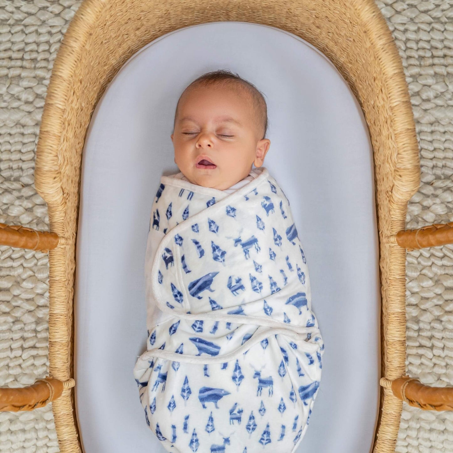 Sleeping baby wrapped in the blue woodland animal swaddle, resting in a woven basket with a white fitted sheet.
