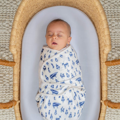 Sleeping baby wrapped in the blue woodland animal swaddle, resting in a woven basket with a white fitted sheet.