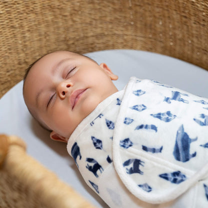 Close-up of sleeping baby swaddled in blue woodland animal print, resting in a woven basket.