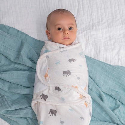 Baby wrapped in the safari animal swaddle lies on a bed with a soft teal muslin beside them.