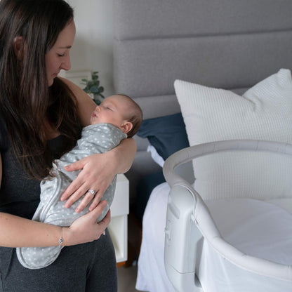 Mother holding her sleeping baby wrapped in the moonlit grey swaddle, standing beside a bedside bassinet.