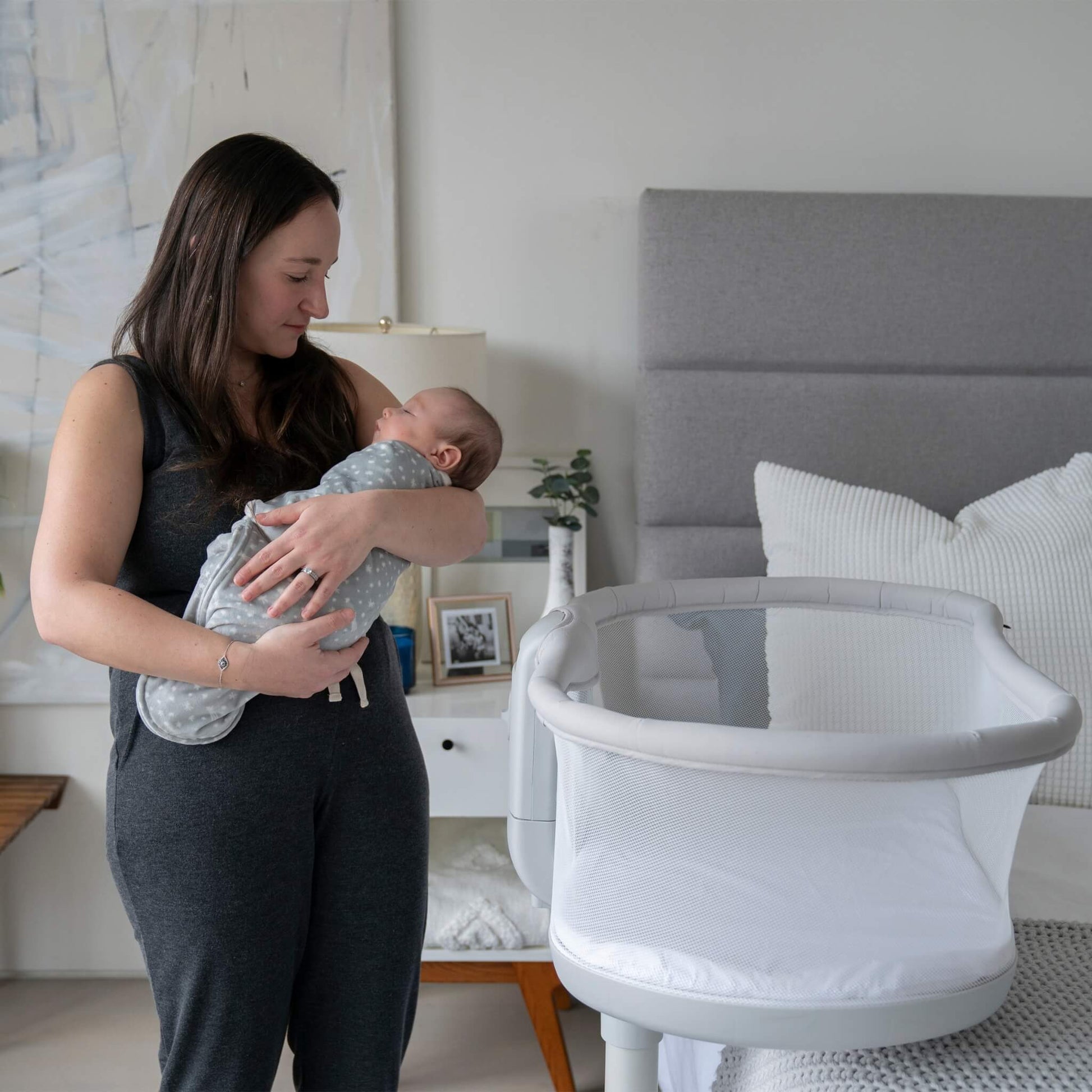 Mother cradling baby in the grey swaddle in a cosy bedroom, looking down lovingly.