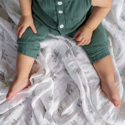 Baby in green romper sitting on soft organic swaddle with zebra and turtle prints by aden + anais, perfect for comfort and play.