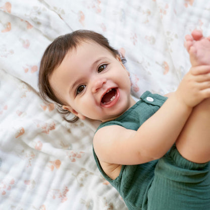 Happy baby lying on the aden + anais organic cotton blanket in Earthly print. Background shows delicate floral and butterfly muslin pattern.