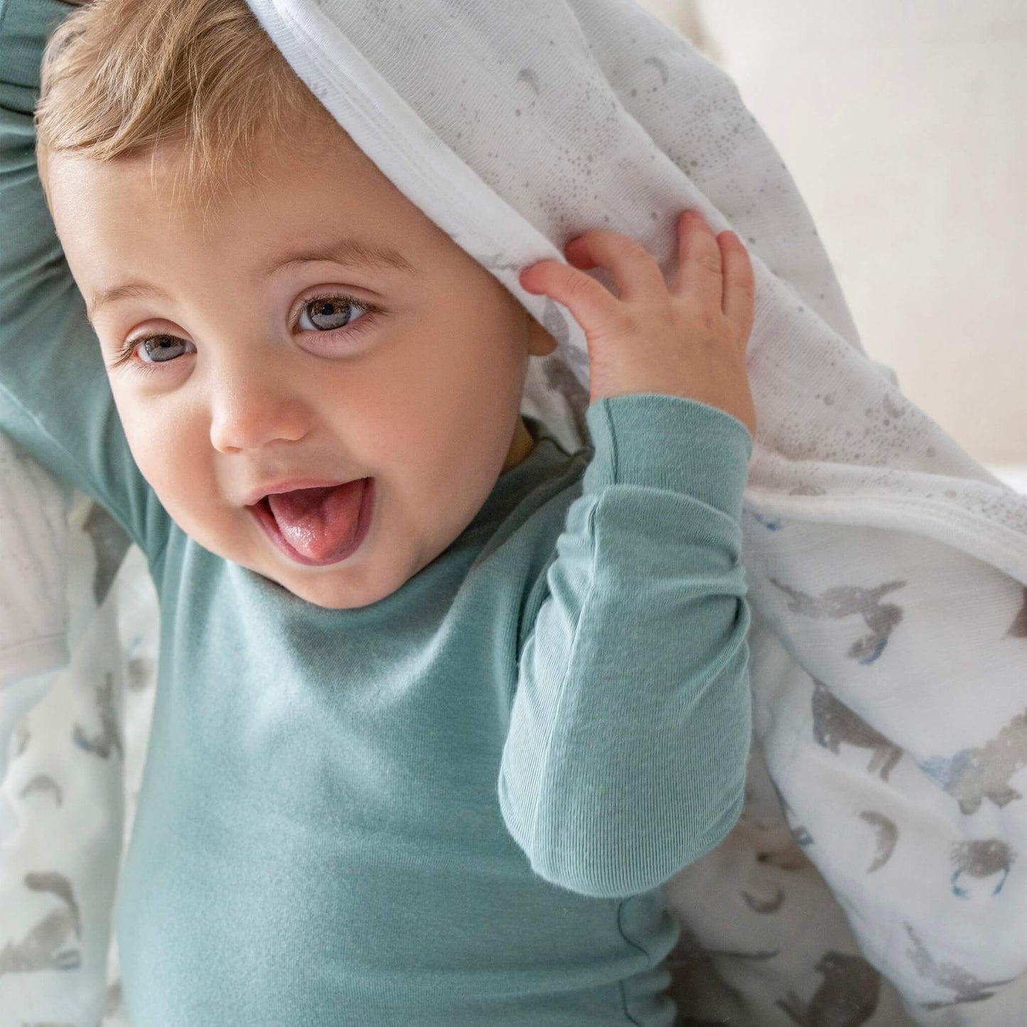 Smiling baby lifting the aden + anais dream blanket over their head. Featuring Map the Stars print in soft grey tones on breathable cotton muslin.