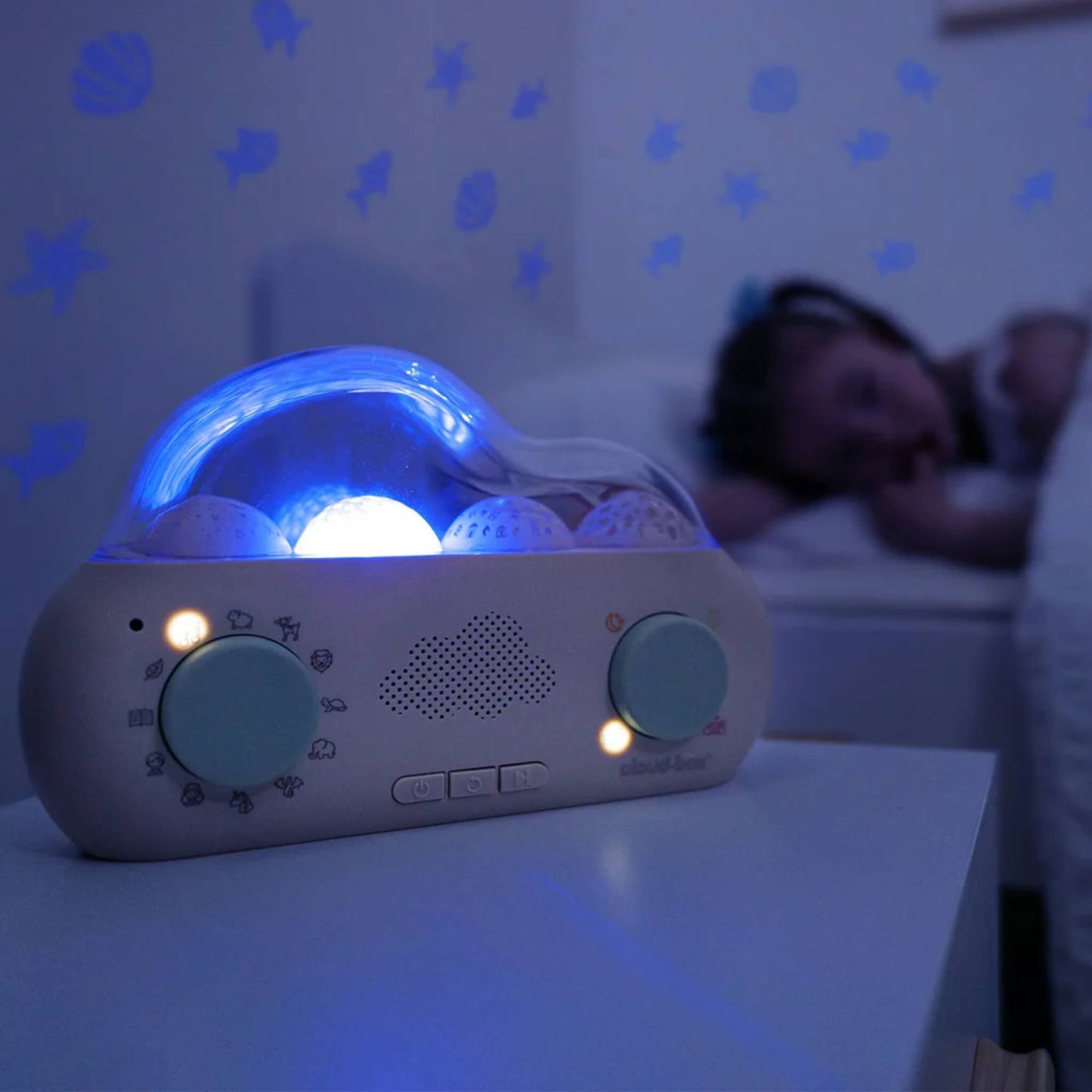 A CloudBox glowing blue on a bedside table, projecting blue stars, leaves and animal shapes onto the wall beside a sleeping child.