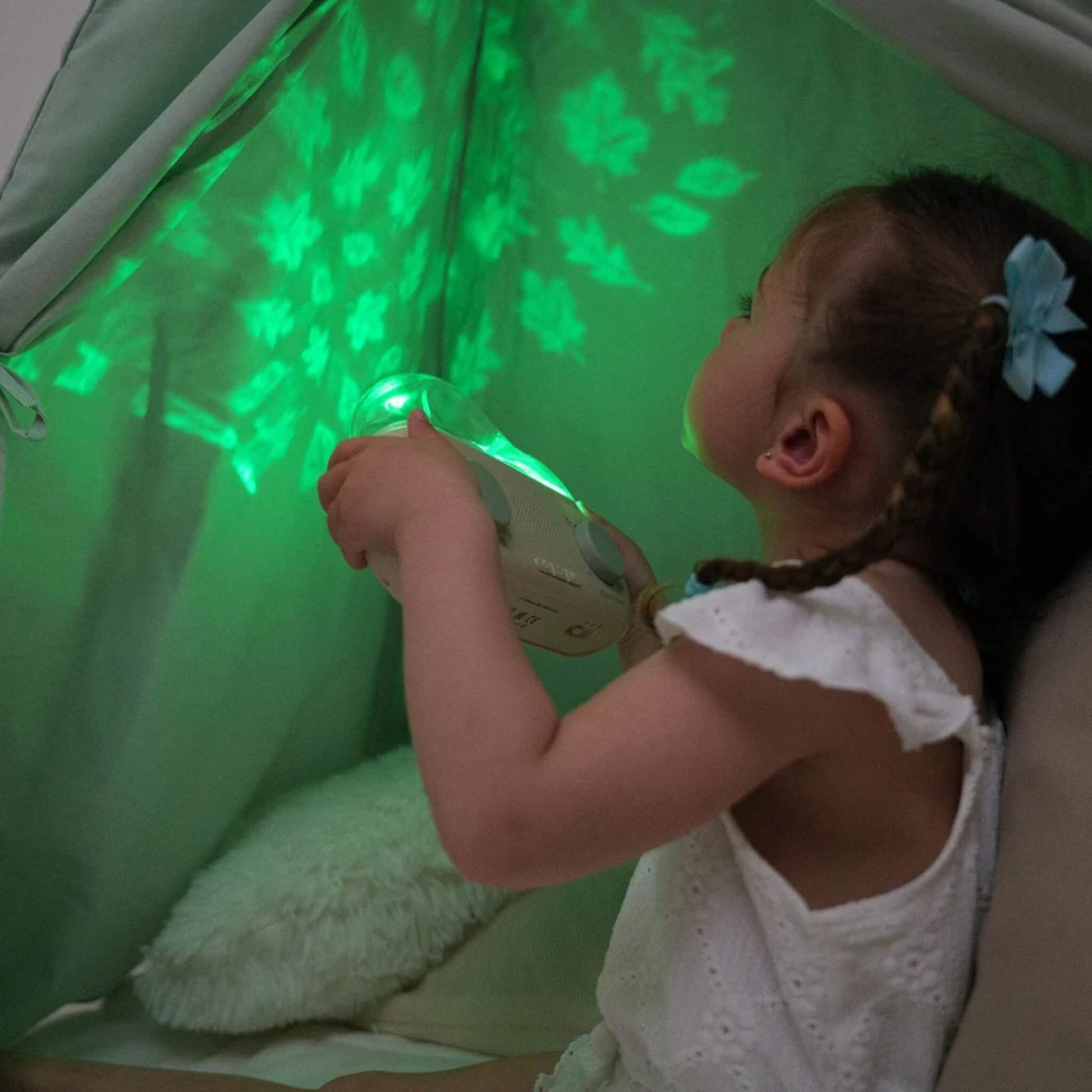 A young girl holding a CloudBox inside a fabric den while bright green leaves, stars and other shapes are projected around her.