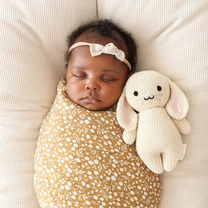 A sleeping newborn baby wrapped in a patterned swaddle lying beside a soft knitted bunny comfort toy.
