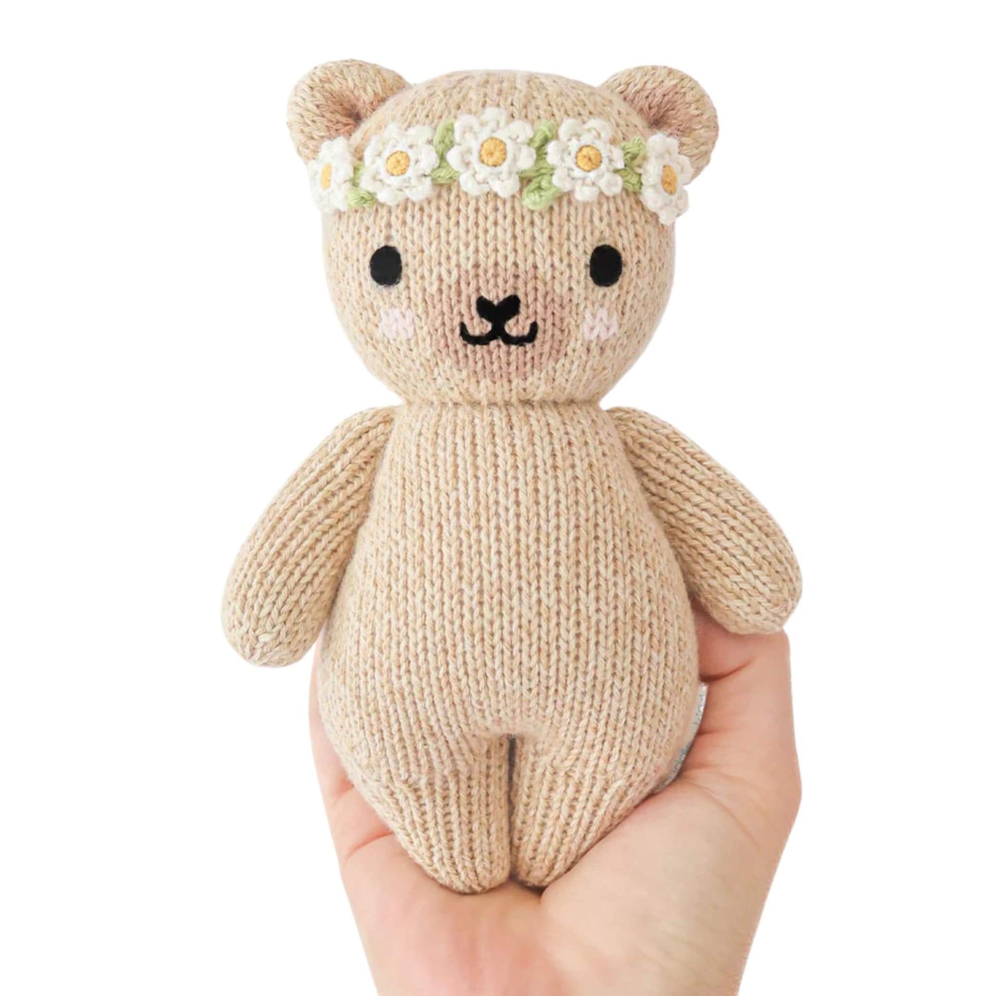 A small knitted teddy bear with a stitched face and white flower crown held in an adult hand to show scale.