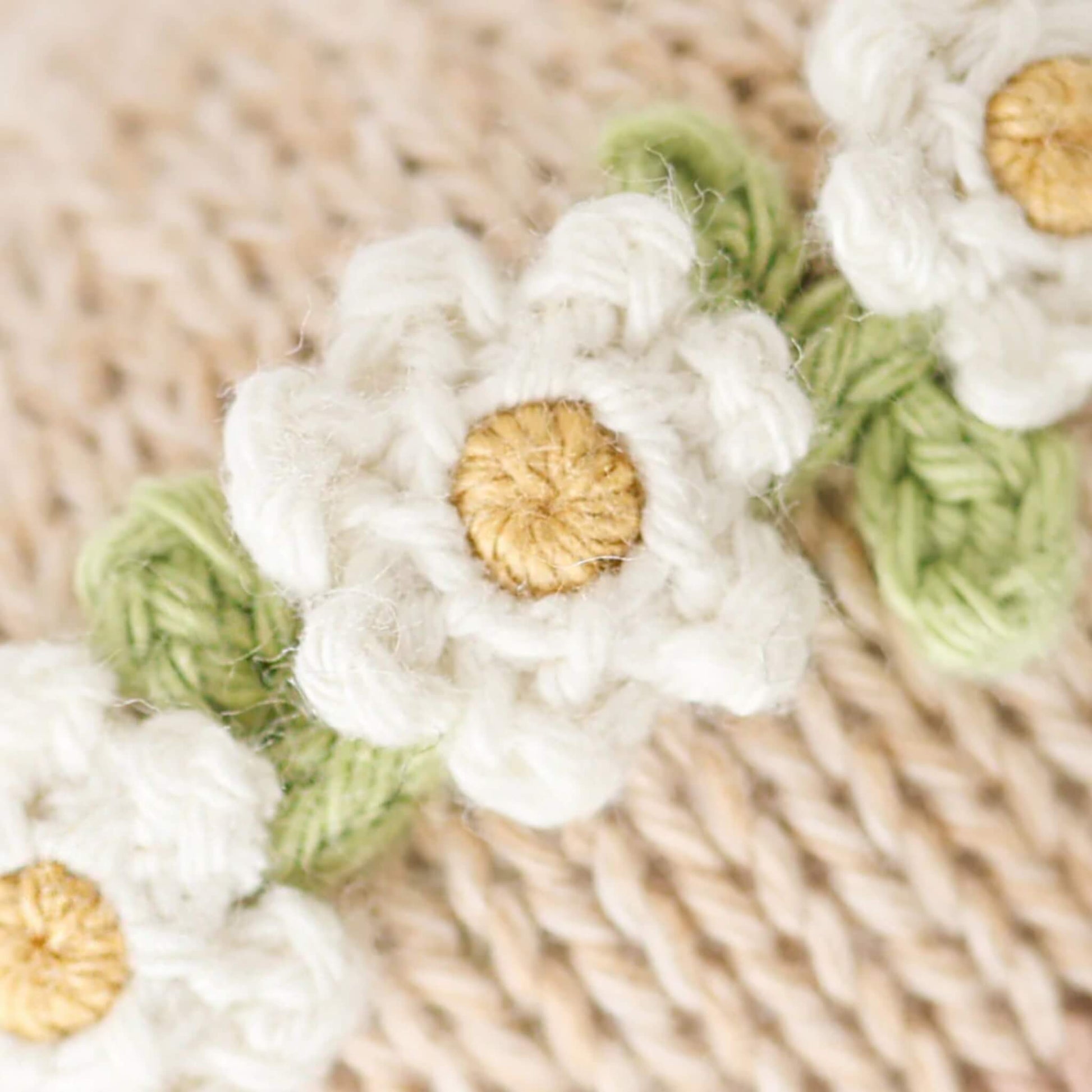 Close-up detail of a knitted daisy flower crown with white petals, yellow centres and green stitching on soft fabric.