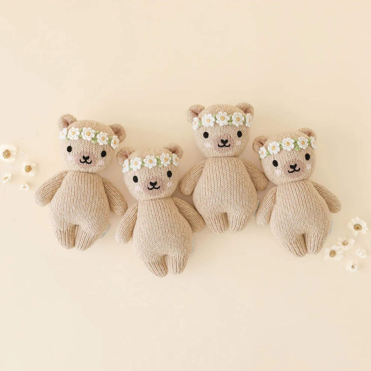 Four knitted teddy bears with stitched faces and flower crowns arranged in a row on a neutral background.