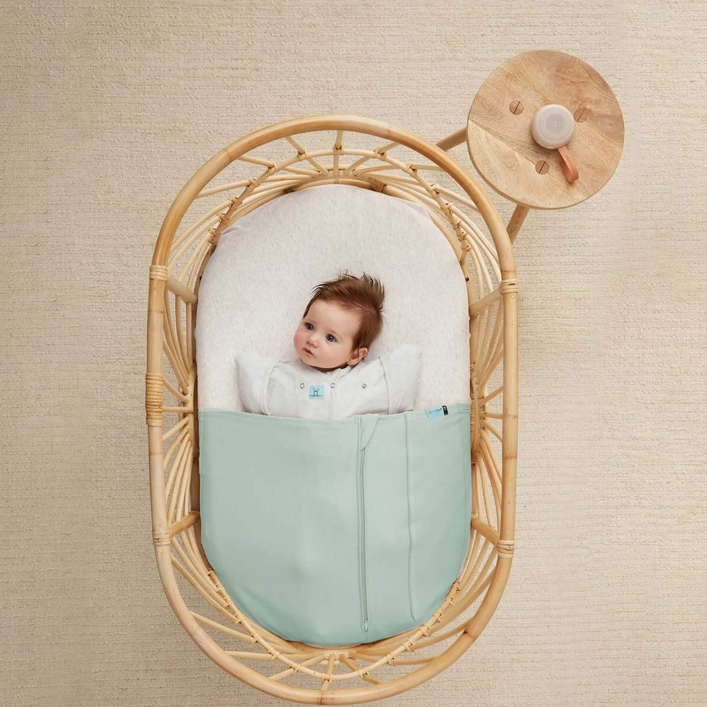 Overhead view of a baby lying in a rattan bassinet wearing a pale green sleep bag, with a small taupe white noise machine placed on a wooden side table nearby.
