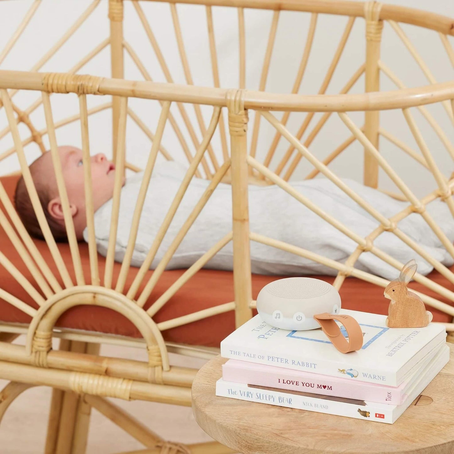 Compact white noise machine resting on top of a stack of children’s books next to a rattan crib, with a resting baby lying inside.