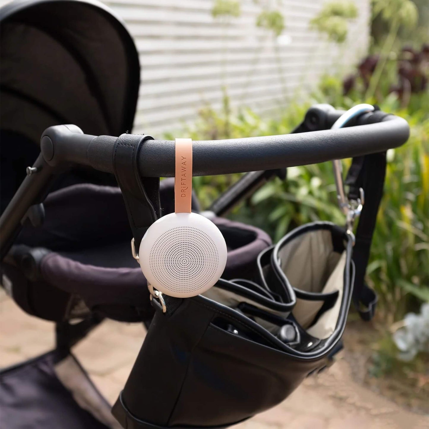 Portable baby sound machine hanging neatly from the handlebar of a black pushchair, secured by its orange strap, with a garden path in the background.