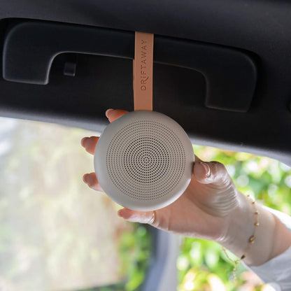Hand holding a compact taupe white noise machine suspended from a car’s overhead handle with its orange strap, showing how it can be used while travelling.