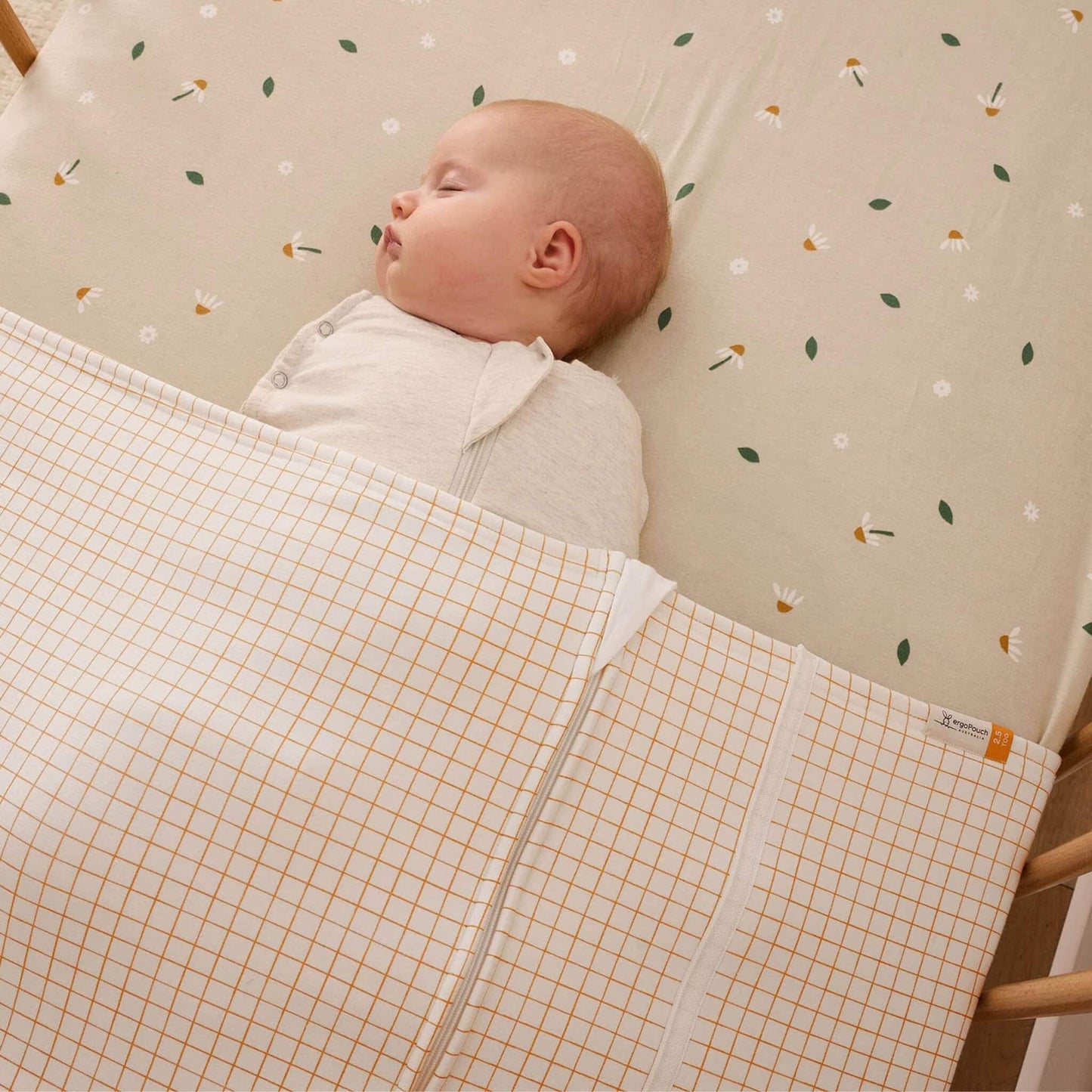 Baby asleep in a cot with a cream and caramel grid blanket tucked over their body.