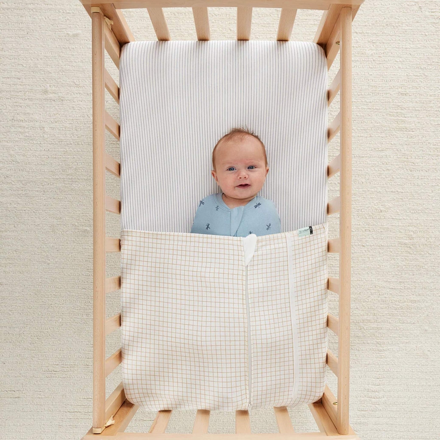 Smiling baby lying in a wooden cot with a caramel grid patterned blanket covering their legs.