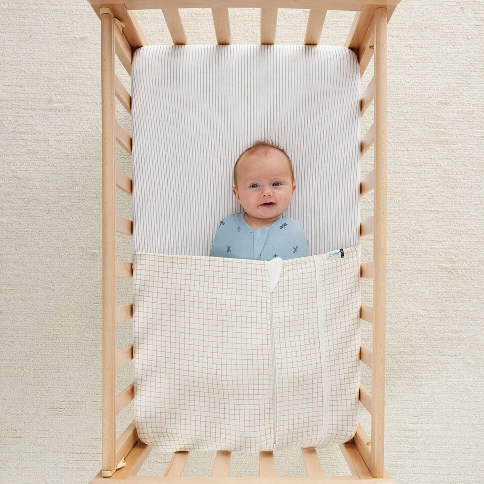 Smiling baby lying in a wooden cot with a caramel grid patterned blanket covering their legs.