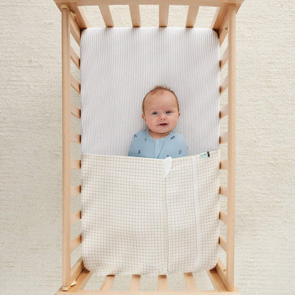 Smiling baby lying in a wooden cot with a caramel grid patterned blanket covering their legs.