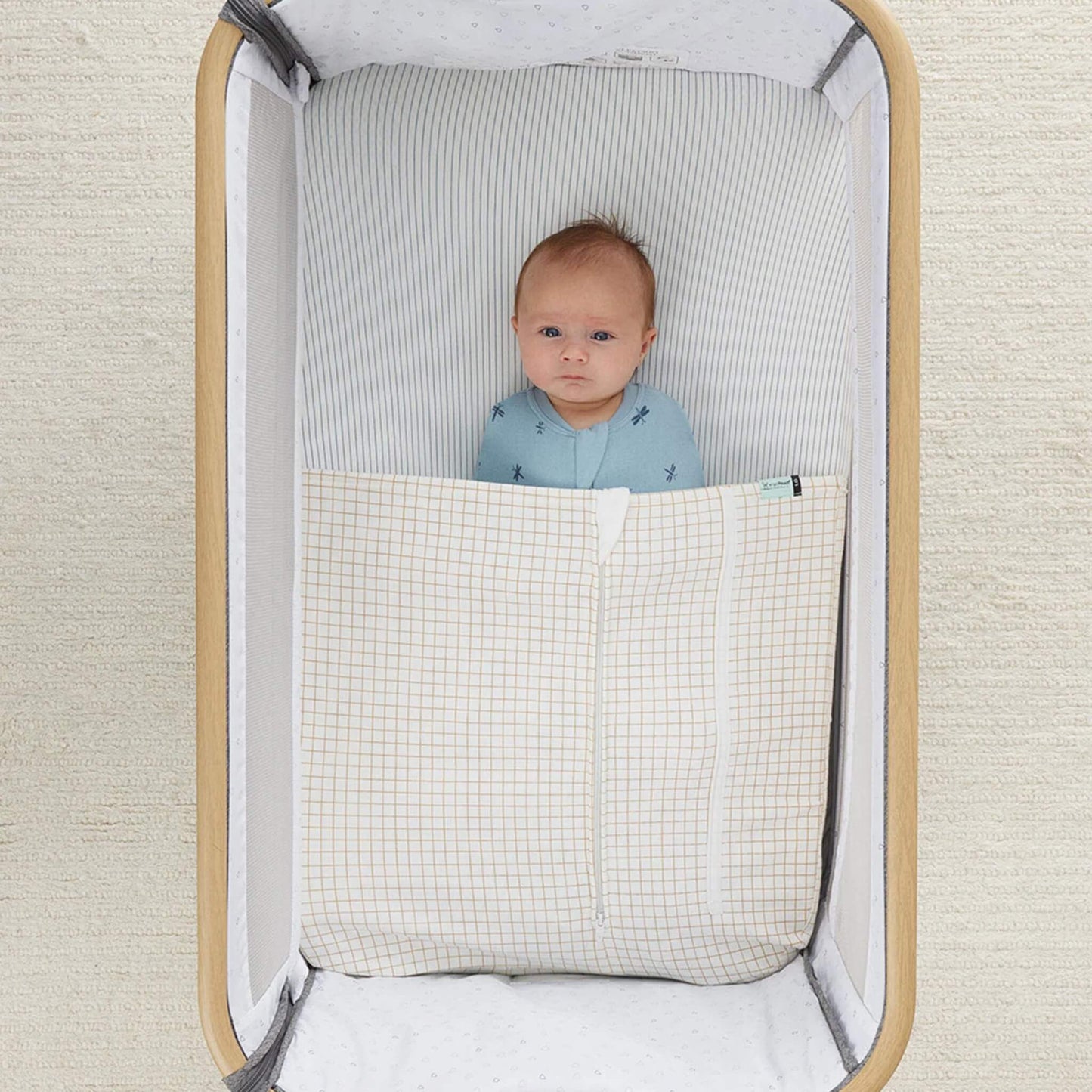 Baby resting in a bedside crib with a cream and caramel grid blanket neatly tucked in.