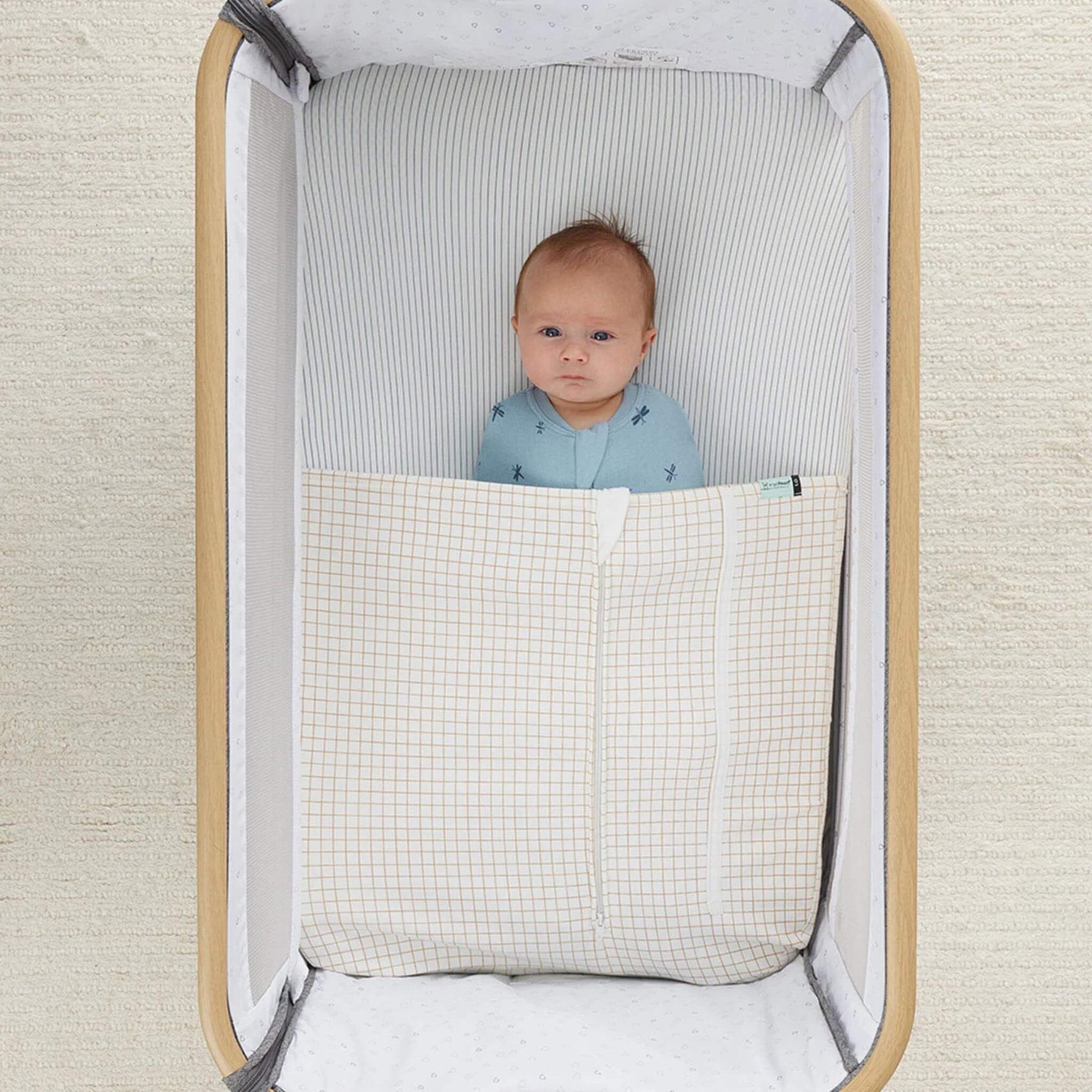 Baby resting in a bedside crib with a cream and caramel grid blanket neatly tucked in.