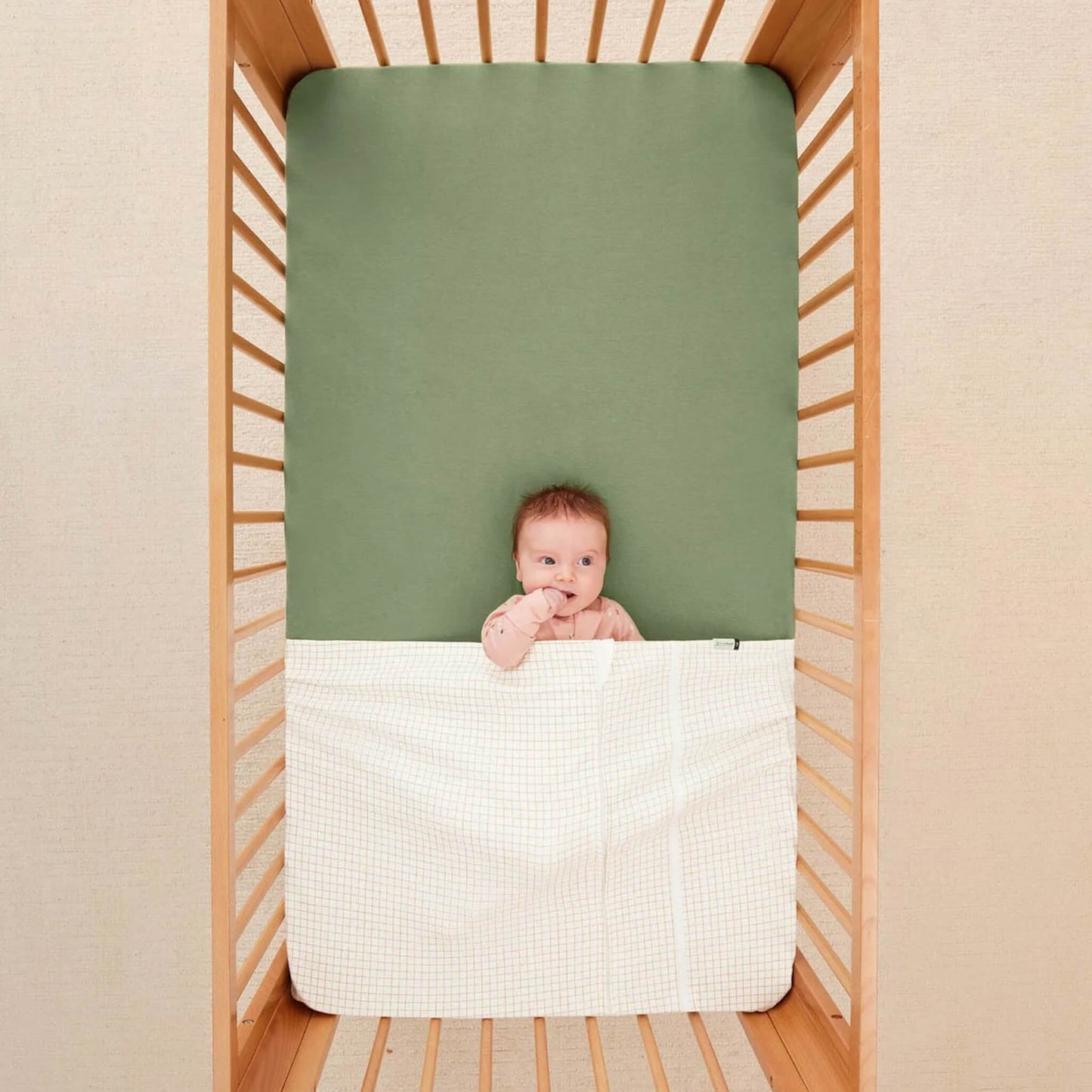 Baby lying in a cot with a green fitted sheet, covered by a caramel grid patterned blanket.