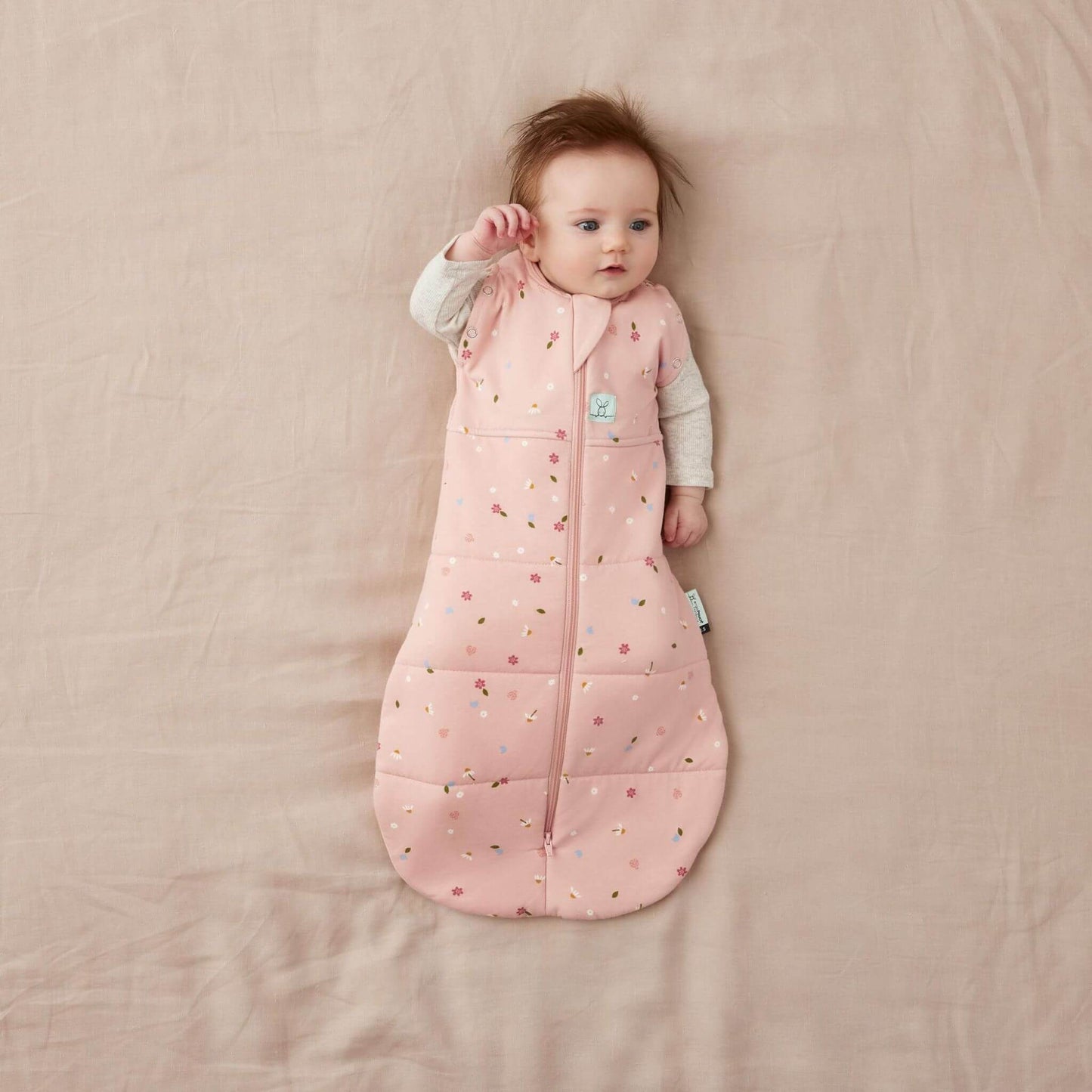 Baby in a Swaddle Bag with a soft pink fabric and a daisies pattern, lying on a neutral bed, arms out and relaxed.