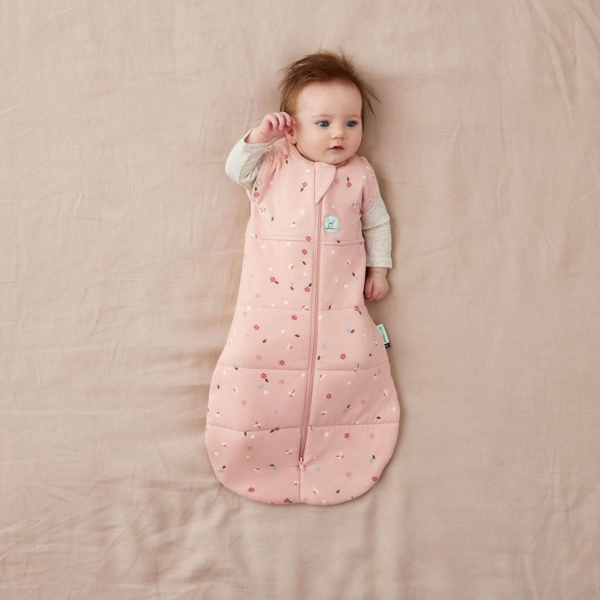 Baby in a Swaddle Bag with a soft pink fabric and a daisies pattern, lying on a neutral bed, arms out and relaxed.