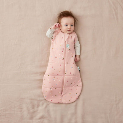 Baby in a Swaddle Bag with a soft pink fabric and a daisies pattern, lying on a neutral bed, arms out and relaxed.