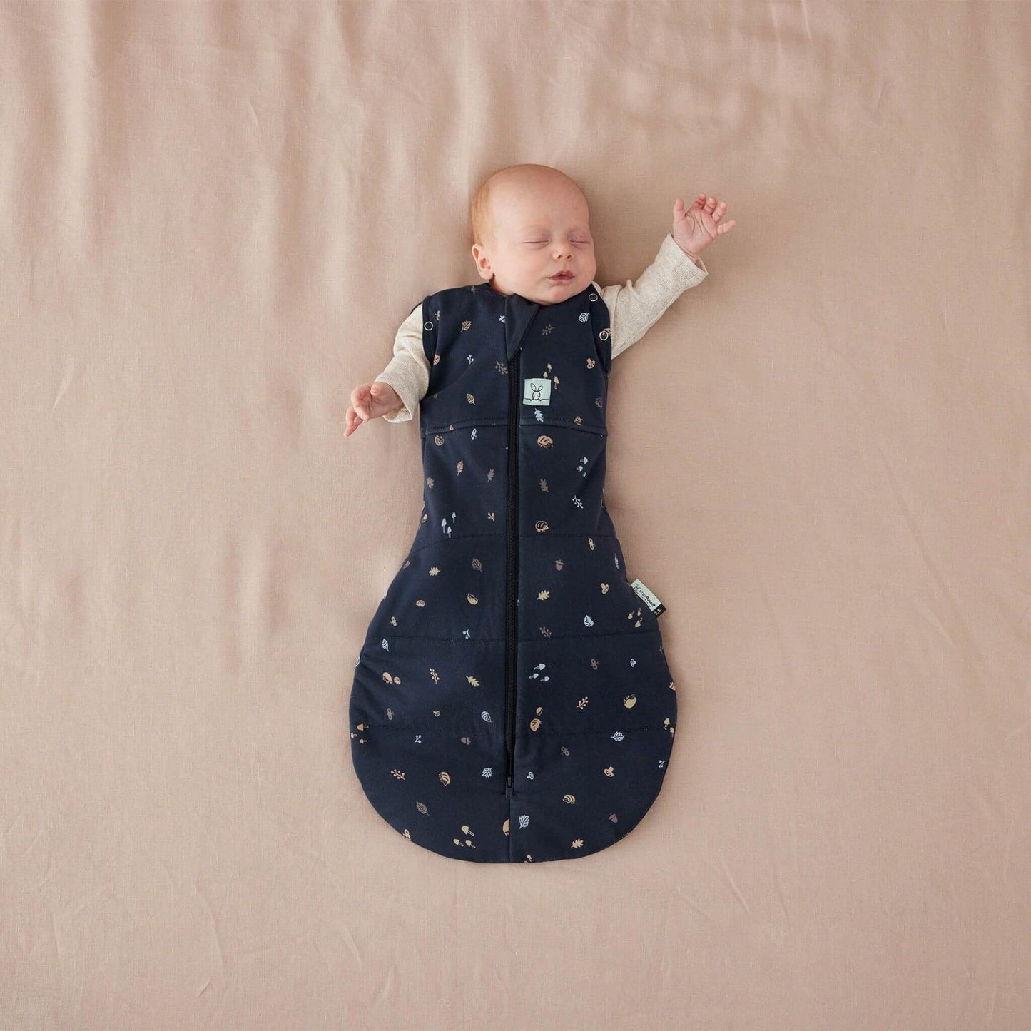 Baby sleeping on a neutral surface in a navy Cocoon Swaddle Bag decorated with woodland-inspired print, both arms free in long-sleeved clothing.
