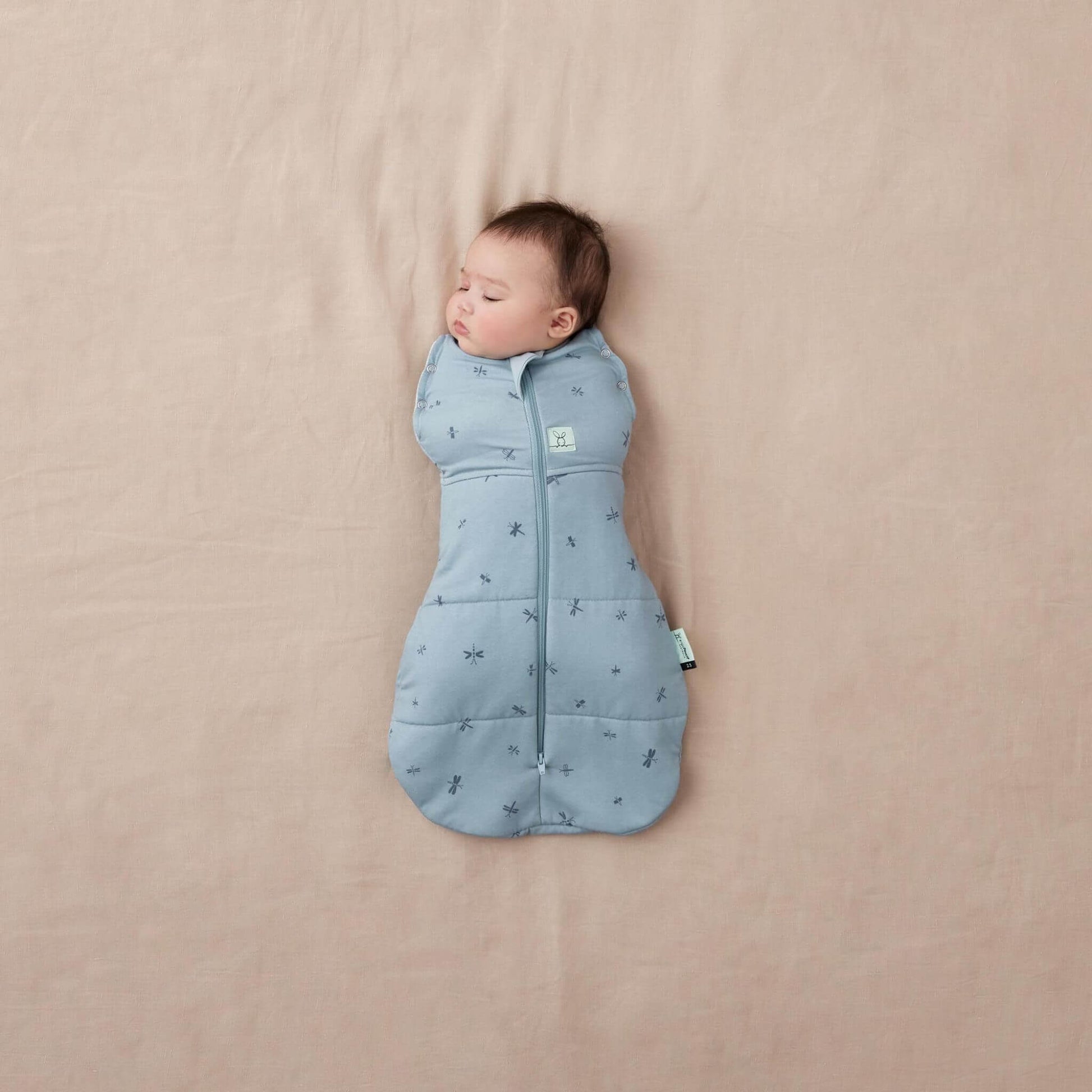 Baby wrapped snugly in a  blue Winter Cocoon Swaddle Bag with dragonflies print, zip-up front, and bell-shaped bottom.
