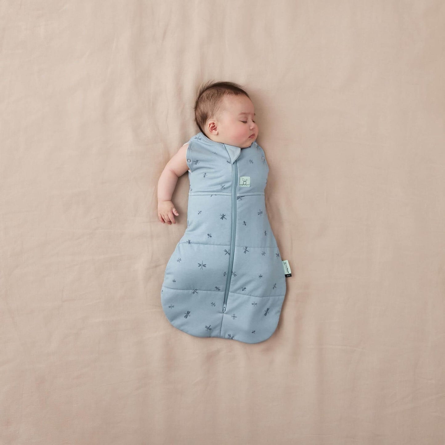 Baby lying comfortably in a blue swaddle Bag with a dragonflies design. Shown with one arm out.