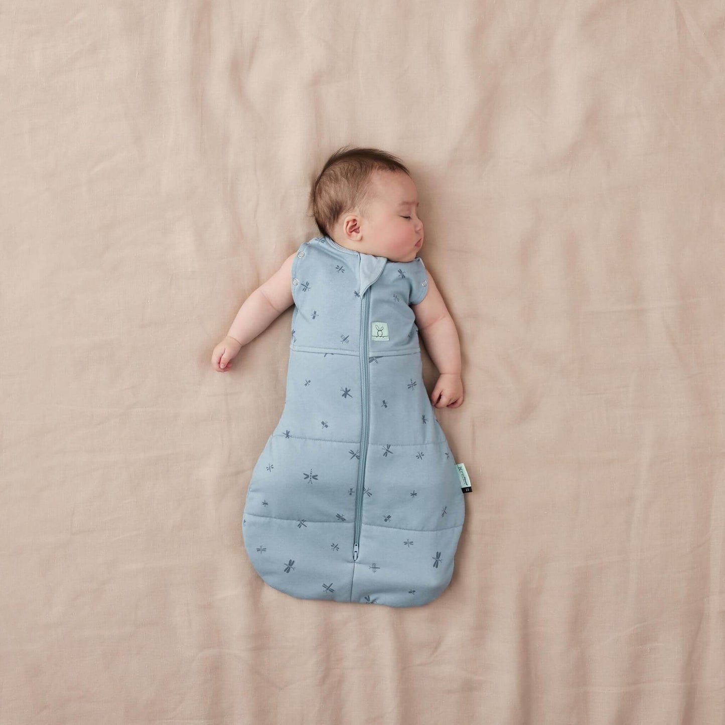 
Baby in a blue cocoon Swaddle Bag with a dragonflies pattern, lying on a neutral bed, both arms out and relaxed.