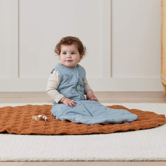 Baby seated on a cosy quilted mat whilst wearing a sleeping Bag, playing with a wooden toy in a bright room.