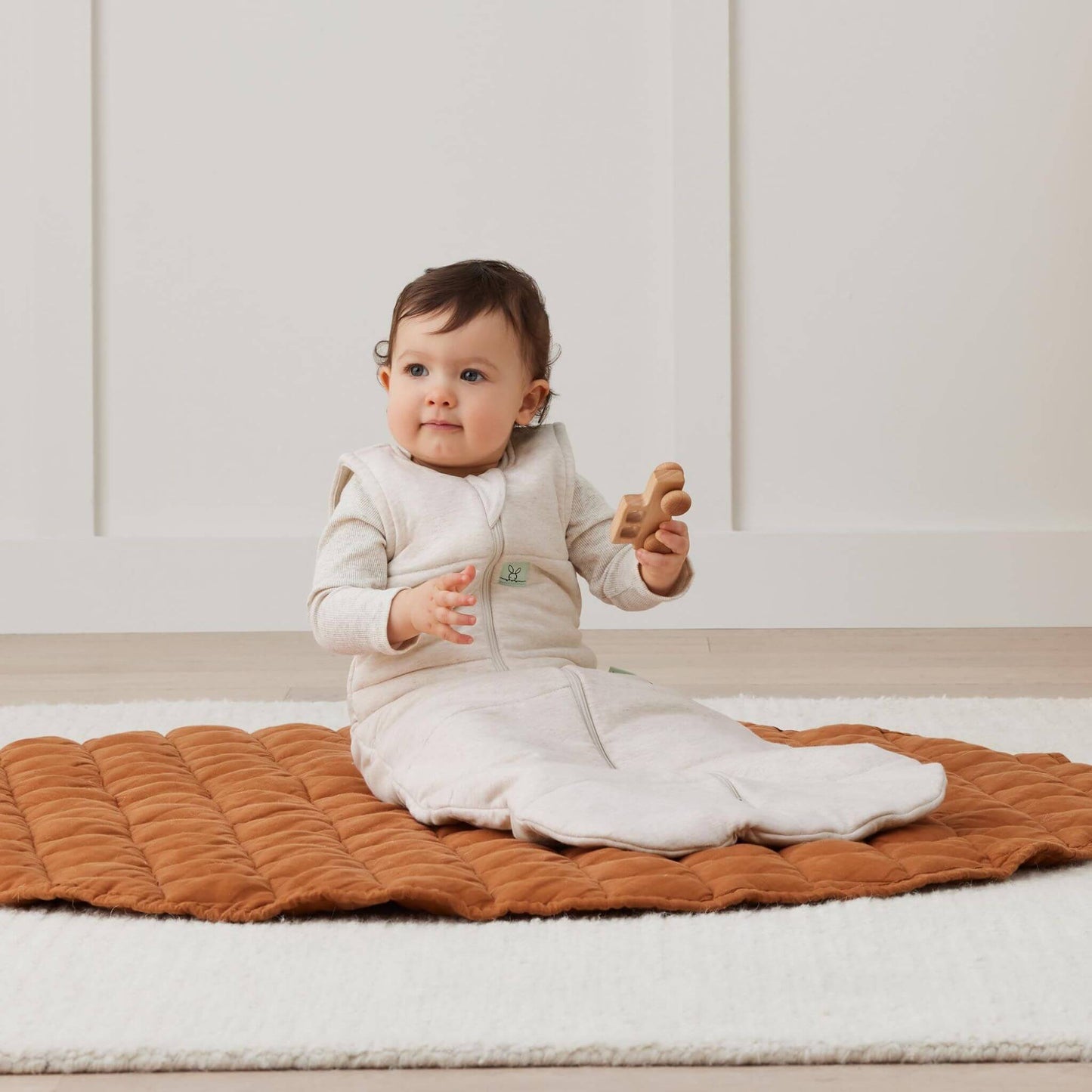 Seated baby wearing an oatmeal beige sleeping bag, while playing with a wooden toy on a cosy quilted mat in a bright room.