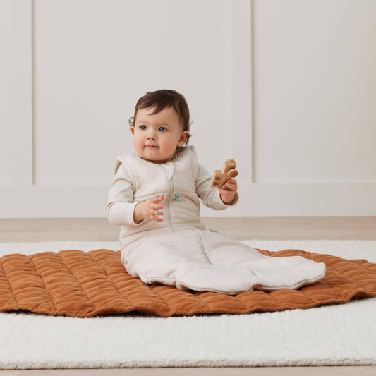 Seated baby wearing an oatmeal beige sleeping bag, while playing with a wooden toy on a cosy quilted mat in a bright room.
