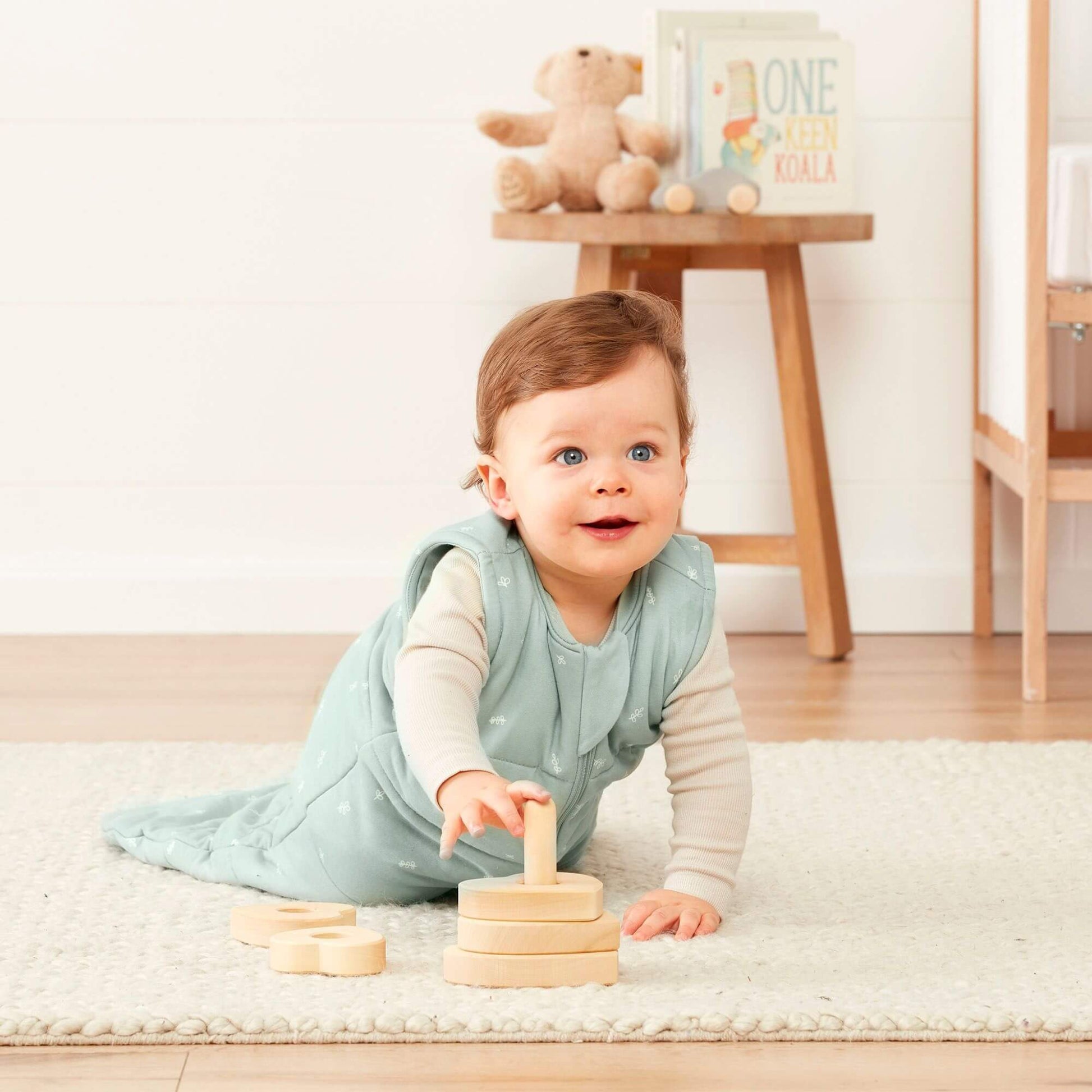 A baby crawls while wearing a sage green sleeping bag, playing with wooden stacking toys in a cosy nursery.
