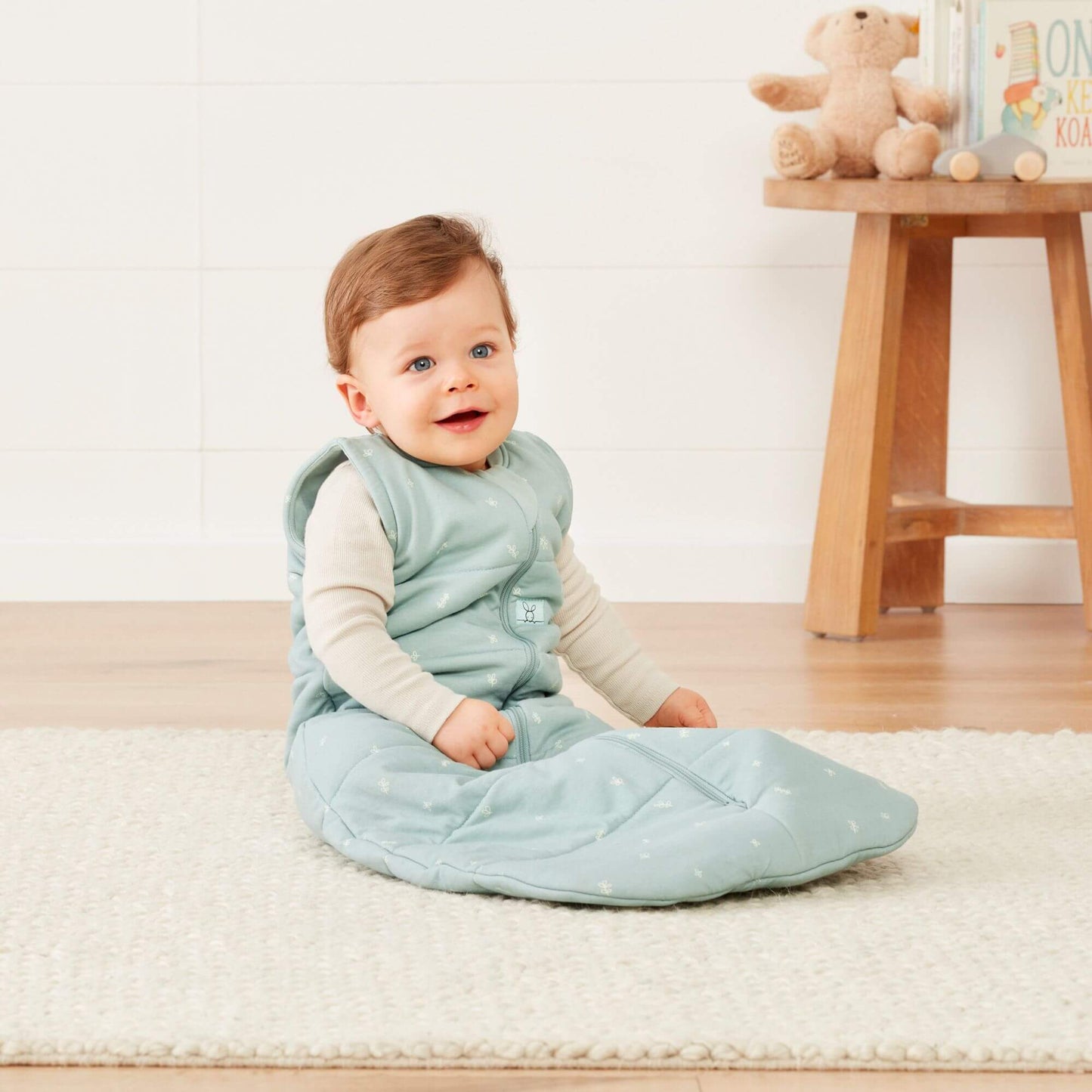 A baby sits on a soft rug wearing a sage green sleeping bag, in a cosy nursery with a wooden stool nearby.