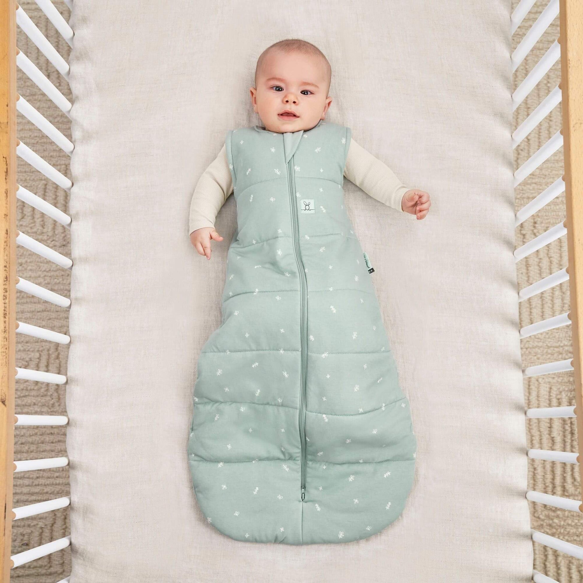 A baby lies in a cot wearing a sage green winter weight sleeping bag, with a central zip and small white patterns.