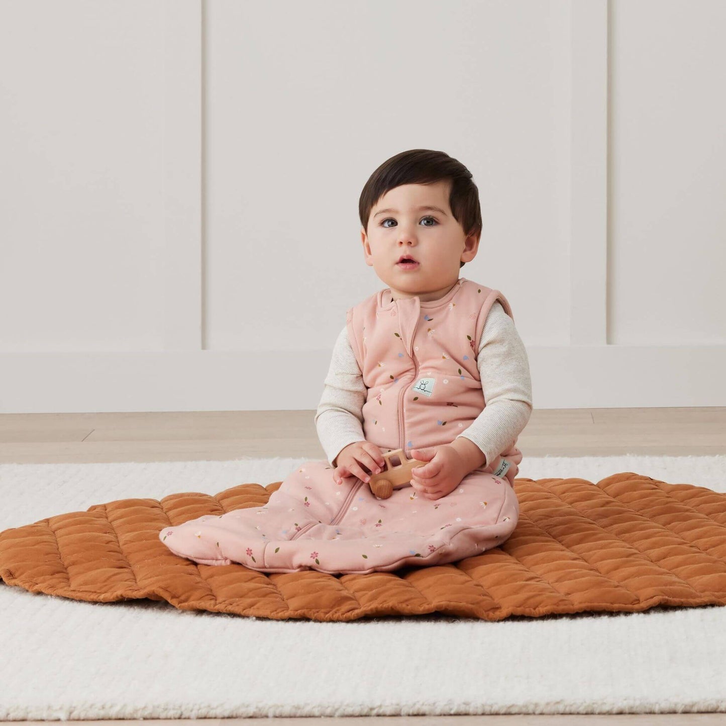 Baby seated whilst wearing a sleeping bag in Daisies print, playing with a wooden toy on a cosy quilted mat in a bright room.