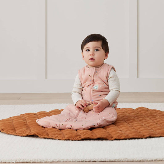 Baby seated whilst wearing a sleeping bag in Daisies print, playing with a wooden toy on a cosy quilted mat in a bright room.